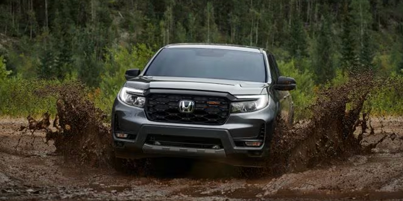 A 2026 Honda Ridgeline driving through muddy terrain, splashing dirt and water.