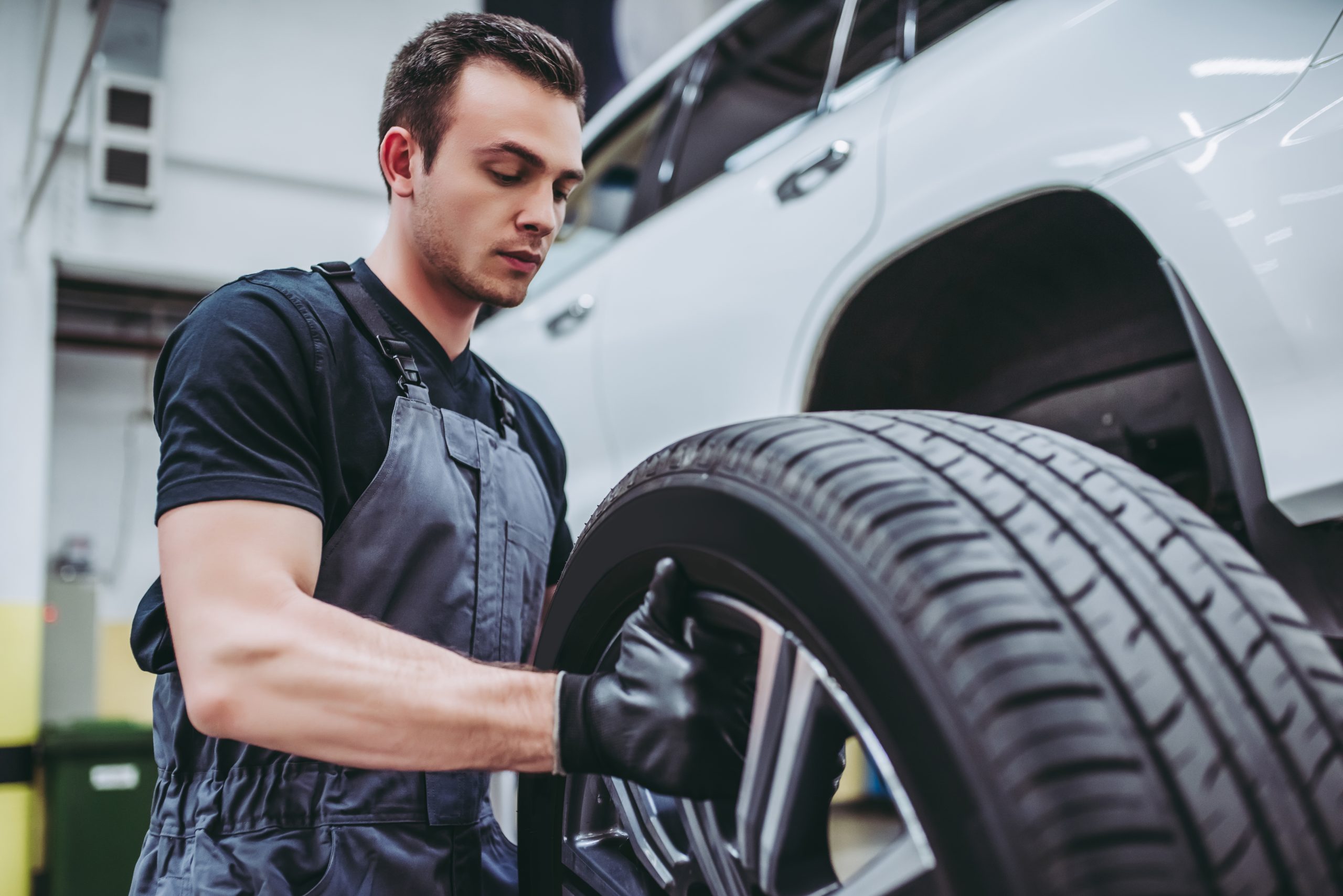 A mechanic is replacing a tire on a vehicle in an auto repair shop. The mechanic is wearing a black shirt, grey overalls, and black gloves while carefully handling the tire. The vehicle in the background is raised, and the workshop is well-lit.