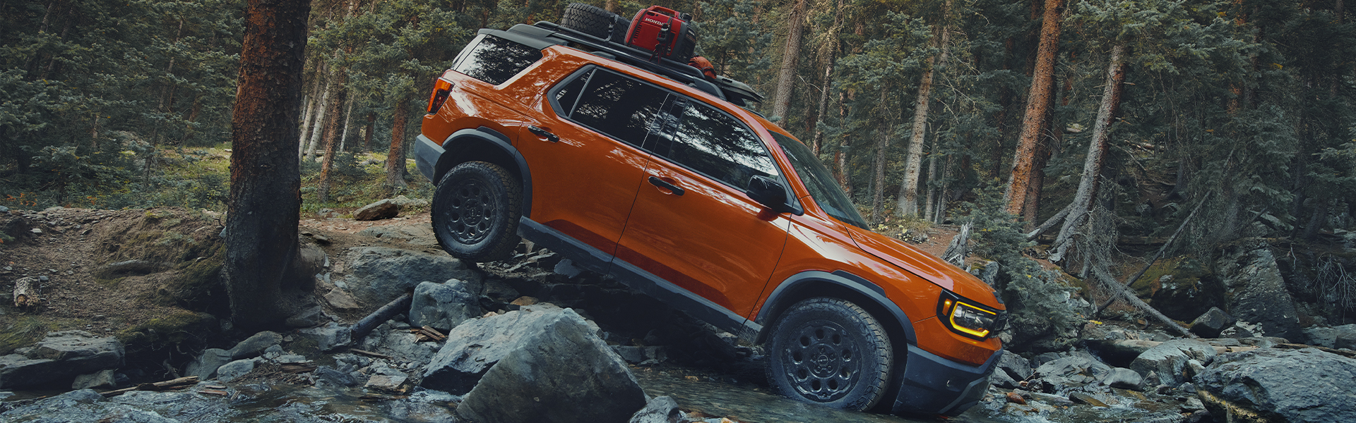 Orange Honda Pilot TrailSport SUV driving over rocks in a pine forest.